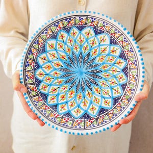 Könnte beinhalten: Eine runde, handbemalte Keramikplatte mit einem lebendigen, symmetrischen Design. Die Platte zeigt ein zentrales blaues Sternmuster, umgeben von komplizierten geometrischen Formen in Blau, Orange, Grün und Rot. Der Rand hat einen gepunkteten Rand.