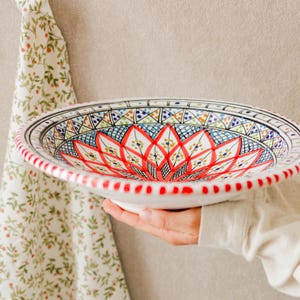 Op de afbeelding: Een witte keramische kom met een levendig bloemmotief in rood, blauw en groen. De rand heeft een stippenpatroon. De kom wordt voor een neutrale achtergrond gehouden, met een bloemmotief doek op de achtergrond.