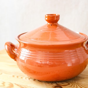 May include: A terracotta cooking pot with a lid, featuring two handles and a small knob on top. The pot is a warm orange color and sits on a wooden surface. The pot is designed for cooking.