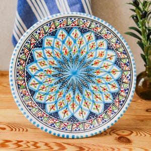 Puede incluir: Un plato redondo de cerámica blanca con un diseño vibrante pintado a mano. El plato presenta un patrón central de estrella azul rodeado de intrincados motivos florales y geométricos en azul, rojo, verde y naranja. El plato se exhibe sobre una superficie de madera.