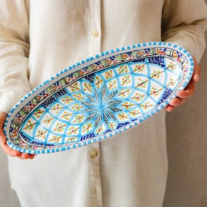 Könnte beinhalten: Eine ovale Servierplatte aus Keramik mit handbemaltem Design. Die Platte hat in der Mitte ein hellblaues Sternmuster, umgeben von blauen, grünen und orangefarbenen geometrischen Formen. Der Rand ist mit blauen Punkten und komplizierten Mustern verziert.