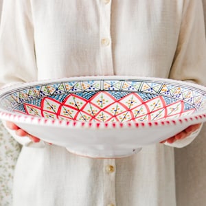 May include: A large, white ceramic serving bowl with a detailed, colourful geometric pattern in red, blue, and green. The rim has a scalloped edge. The bowl is held by a person wearing a white shirt.