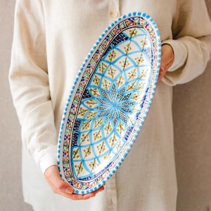 Könnte beinhalten: Eine ovale Servierplatte aus Keramik mit handbemaltem Design. Die Platte hat ein zentrales blaues Sternmuster, umgeben von geometrischen und floralen Motiven in Blau, Orange und Grün. Der Rand ist mit blauen Punkten verziert.
