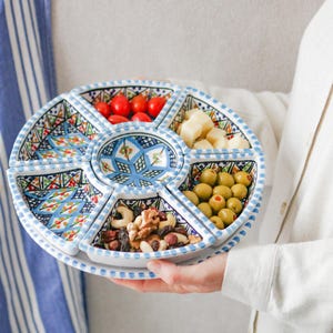 May include: A circular serving platter with a blue and white patterned design, filled with olives, nuts, cheese, and tomatoes. The dish is held by a person wearing a white shirt.