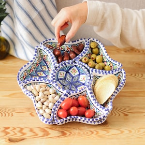 Könnte beinhalten: Ein dekoratives Serviertablett mit mehreren Fächern und einem blau-weißen Blumendesign. Es ist gefüllt mit Oliven, Datteln, Pistazien, Tomaten und Crackern. Eine Hand greift nach einer Dattel. Das Tablett steht auf einer Holzoberfläche.