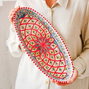 Könnte beinhalten: Eine ovale Servierplatte aus Keramik mit einem lebendigen, komplizierten Blumendesign. Die Platte hat ein rotes zentrales Motiv mit grünen, blauen und weißen Akzenten. Der Rand ist mit roten Punkten verziert. Ein dekoratives Stück zum Servieren von Speisen.