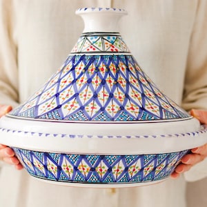 Peut inclure: Un tajine en céramique blanc et bleu avec un couvercle conique. Le couvercle et la base sont décorés d'un motif géométrique bleu, rouge, vert et orange. Le tajine est tenu par une personne.
