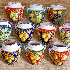Puede incluir: Una colección de diez jarrones de cerámica con asas, cada uno con diseños pintados a mano. Los jarrones muestran colores vibrantes como el rojo, azul, amarillo y verde, con patrones de frutas y flores. Los jarrones tienen un borde blanco y un interior de terracota.