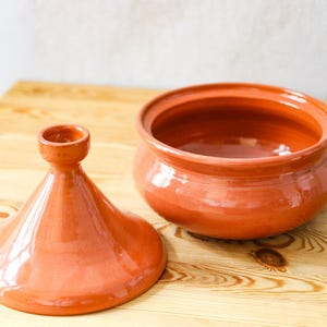 Può includere: Una pentola tagine in terracotta aperta, con il suo coperchio conico accanto. La pentola ha una base arrotondata e rivela l'interno. Il tagine è su una superficie di legno chiaro.