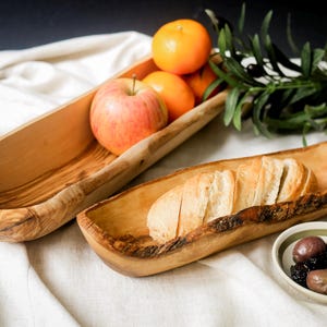May include: Two wooden serving bowls, one holding apples and oranges, the other with sliced bread. A small bowl of olives is next to the bread bowl. The bowls are on a white cloth.