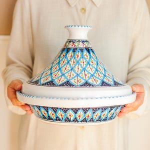 May include: A white ceramic tagine pot with a conical lid, decorated with a blue, green, and red geometric pattern. The pot is held by a person, highlighting its intricate design and traditional shape. The tagine is a classic cooking vessel.
