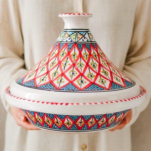 Peut inclure: Un tajine en céramique blanche avec un couvercle conique, orné d'un motif vibrant de losanges rouges, d'accents bleus et de détails floraux verts et jaunes. Le pot est tenu par une personne portant une chemise crème.