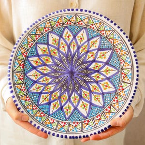 May include: A decorative ceramic plate with a vibrant floral design. The plate features a central purple flower surrounded by blue, green, orange, and red geometric patterns. The rim is adorned with small blue dots.
