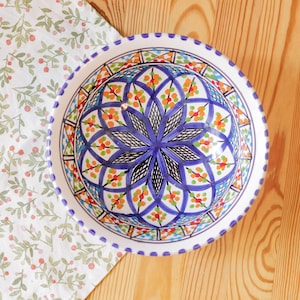 Op de afbeelding: Een ronde, handbeschilderde keramische kom met een levendig bloemenontwerp. De kom heeft een centraal blauw sterpatroon, omringd door oranje, groene en blauwe bloemmotieven. De rand heeft een gestippelde rand. De kom staat op een houten oppervlak.