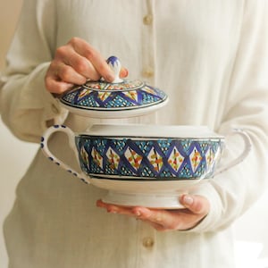 Può includere: Una zuppiera in ceramica bianca con coperchio, caratterizzata da un vivace motivo geometrico blu, giallo e arancione. La zuppiera ha due manici e una base a piedistallo. Il coperchio viene sollevato, rivelando l'interno.