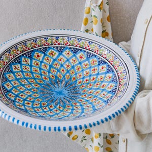 May include: A large, round ceramic bowl with a white rim and a detailed pattern. The bowl has a blue, red, yellow, and green floral design in the centre. The rim features a blue dotted pattern. The bowl's diameter is approximately 30 cm.