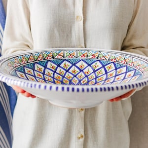 Puede incluir: Un gran cuenco de cerámica blanca con un detallado y colorido patrón geométrico en azul, verde, rojo y amarillo. El borde tiene un borde festoneado con detalles azules. Una pieza decorativa para servir alimentos.