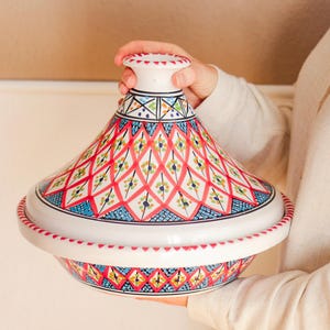 Op de afbeelding: Een witte keramische tajinepot met een conisch deksel, met een levendig handgeschilderd ontwerp. Het patroon bevat rode, blauwe en gele geometrische vormen. De pot wordt door een persoon vastgehouden.