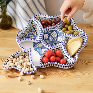 May include: A white ceramic serving dish with a blue and turquoise floral design, filled with olives, tomatoes, pistachios, dates, and crackers. The dish is on a wooden table.