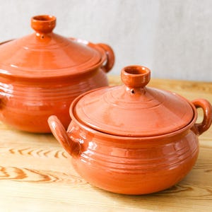 Puede incluir: Dos ollas de cocina de terracota con tapas. Las ollas son de color naranja cálido y tienen asas en los lados. Están sobre una superficie de madera de color claro.