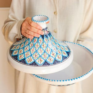 Può includere: Un tajine in ceramica bianca con coperchio conico e base larga. Il coperchio è decorato con un motivo geometrico blu e bianco, e la base ha un bordo a pois blu. Un tradizionale recipiente da cucina marocchino.