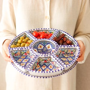 Könnte beinhalten: Ein runder, geteilter Servierteller mit blauem und weißem geometrischem Design, gefüllt mit Oliven, Tomaten und Datteln. Der Teller wird von einer Person gehalten, die ein cremefarbenes Hemd trägt.