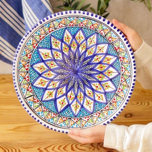 Könnte beinhalten: Eine runde, handbemalte Keramikplatte mit einem lebendigen Blumenmuster. Die Platte hat ein zentrales blaues Blumenmotiv, umgeben von komplizierten Mustern in Blau, Orange, Grün und Rot. Der Rand ist mit einem gepunkteten Rand verziert. Die Platte wird von zwei Händen gehalten. Durchmesser: ca. 25 cm.