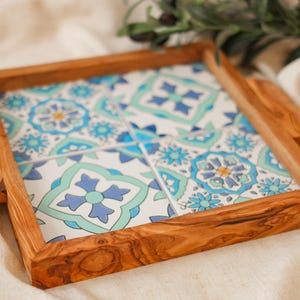 Könnte beinhalten: Quadratisches Serviertablett aus Holz mit dekorativem Fliesen-Einsatz. Das Fliesendesign zeigt ein sich wiederholendes Muster aus blauen und türkisfarbenen Blumenmotiven auf weißem Grund. Das Tablett hat geschnitzte Griffe an jeder Seite.