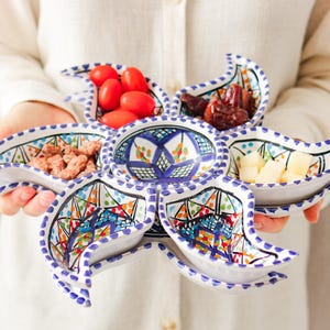 Op de afbeelding: Een witte keramische serveerschaal met een bloemenpatroon, gevuld met diverse snacks. De schaal heeft een centrale kom en zes bloemblad-vormige compartimenten, elk met verschillende voedingsmiddelen zoals tomaten en noten. De schaal wordt door een persoon vastgehouden.