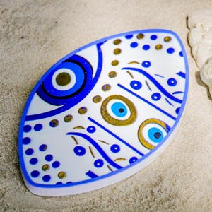 Op de afbeelding: Witte, ovale decoratieve schaal met een blauwe rand. De schaal heeft een patroon van blauwe en gouden oogontwerpen en stippen. De schaal is waarschijnlijk gemaakt van keramiek of een vergelijkbaar materiaal.