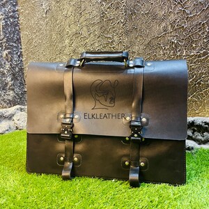May include: Black leather briefcase with a minimalist line drawing of a face and the text "ELKLEATHER" on the front. The briefcase has two buckles on each side and a top handle.