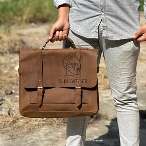 May include: A brown leather briefcase with a flap closure and two buckle straps. The bag has a top handle and a shoulder strap. The text "ELKLEATHER" is visible on the front of the bag. The bag is held by a person.