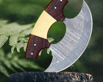 May include: A large, curved, stainless steel knife with a wooden handle. The blade has a unique, patterned finish. The knife is resting on a wooden stump.
