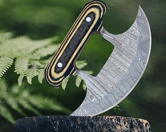 May include: A Damascus steel Ulu knife with a curved blade and a wooden handle. The handle is made of black and tan wood with two white rivets. The knife is displayed on a tree stump against a blurred green background.