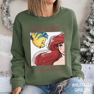 May include: Military green sweatshirt with a graphic of Ariel and Flounder from The Little Mermaid, and the text "BUT DADDY I Love Him" in red. The sweatshirt is a solid colour and has a crew neck.