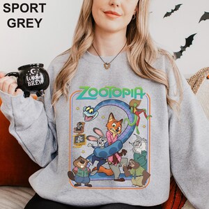 May include: Sport grey sweatshirt featuring a colorful graphic from the movie Zootopia. The design includes characters like Judy Hopps, Nick Wilde, and others, with the word "Zootopia" in a retro font. The sweatshirt is a crew neck style.