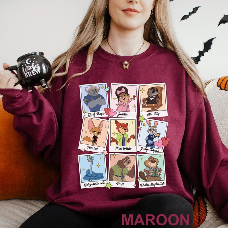 Pu&ograve; includere: Felpa bordeaux con una griglia di ritratti di personaggi dei cartoni animati, tra cui Chief Bogo, Nick Wilde e Judy Hopps. La parola "MAROON" &egrave; stampata in basso. Una tazza nera con "Witches Brew" &egrave; tenuta in mano.