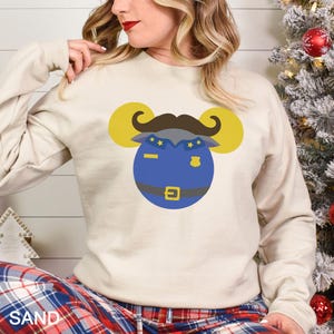 May include: A sand-colored sweatshirt featuring a cartoon police officer design. The design includes a blue police uniform, a brown mustache, and yellow mouse ears. The sweatshirt is long-sleeved and has a crew neck.