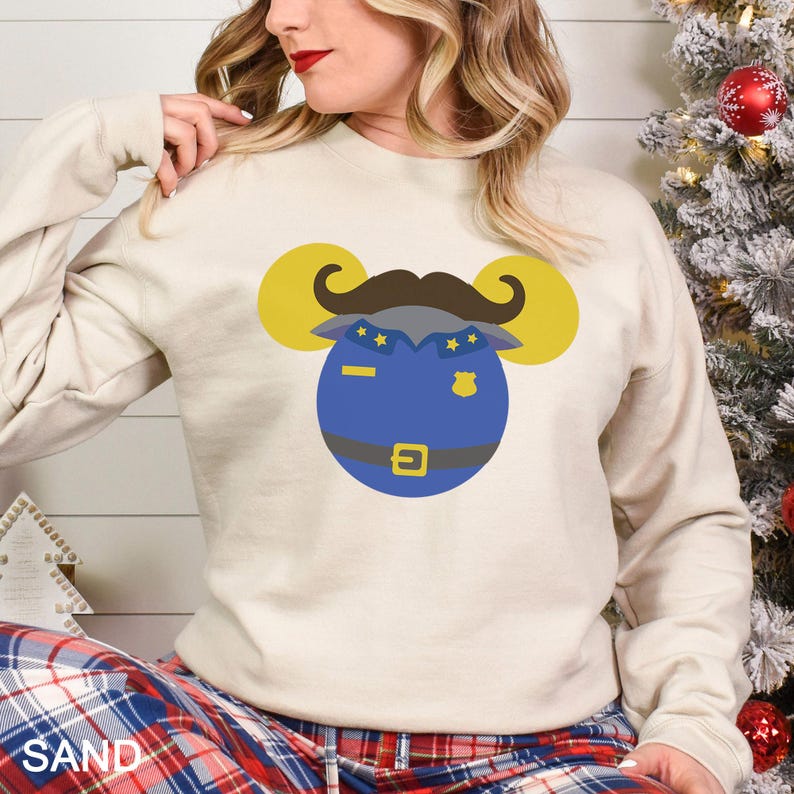 May include: A sand-colored sweatshirt featuring a cartoon police officer design. The design includes a blue police uniform, a brown mustache, and yellow mouse ears. The sweatshirt is long-sleeved and has a crew neck.
