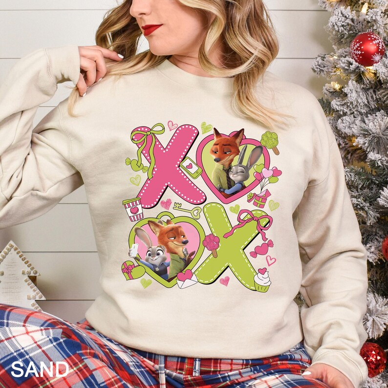 Peut inclure: Un sweat-shirt couleur sable avec un motif rose et vert repr&eacute;sentant les lettres "X" et "O". Le motif comprend des images de dessins anim&eacute;s d'un renard et d'un lapin &agrave; l'int&eacute;rieur de coeurs, ainsi que d'autres petits graphismes.