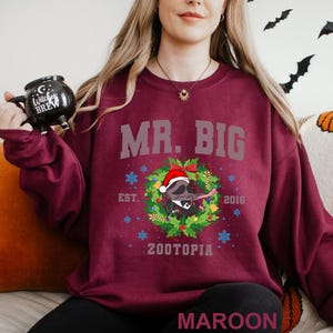 Pu&ograve; includere: Felpa bordeaux con la scritta "MR. BIG" sopra un procione con un cappello di Babbo Natale all'interno di una ghirlanda. La felpa ha anche la scritta "EST. 2016" e "ZOOTOPIA". Una tazza nera con "Witches Brew" &egrave; tenuta nella mano sinistra.