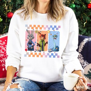 May include: White sweatshirt featuring three cartoon characters: a rabbit in a police uniform, a fox in a green jacket, and a snake. The design is framed with a checkered pattern in orange, pink, and blue.