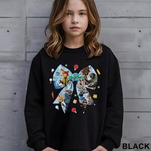 May include: Black sweatshirt featuring a large bow design with characters from the animated film "Zootopia." The bow is decorated with cartoon images of animals and colorful accents. The word "BLACK" is printed at the bottom.