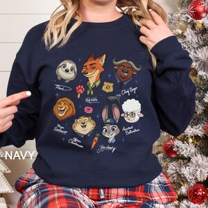 K&ouml;nnte beinhalten: Marineblaues Sweatshirt mit Cartoon-Illustrationen von Charakteren aus dem Film Zootopia, darunter Flash, Nick Wilde, Chief Bogo und Judy Hopps. Das Design zeigt auch andere Charaktere. Das Wort "NAVY" ist unten links aufgedruckt.