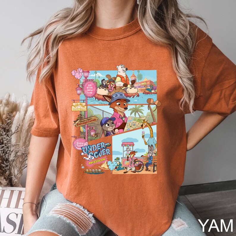 May include: A burnt orange t-shirt featuring a multi-panel graphic print of animated animal characters in a theme park setting. The design includes various scenes with characters, park signage, and the text Undercover Mission in blue and white lettering.