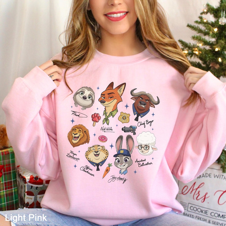 K&ouml;nnte beinhalten: Hellrosa Sweatshirt mit Cartoon-Illustrationen von Zootopia-Charakteren, darunter Nick Wilde und Chief Bogo. Das Design umfasst Charakternamen und verschiedene Illustrationen. Das Sweatshirt hat eine hellrosa Farbe.