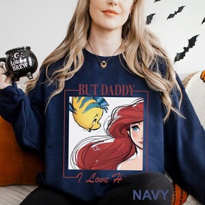 May include: Navy blue sweatshirt with a graphic of Ariel and Flounder from The Little Mermaid, with the text "BUT DADDY - I Love Her" in white. The word "NAVY" is printed at the bottom. The sweatshirt is a casual style.