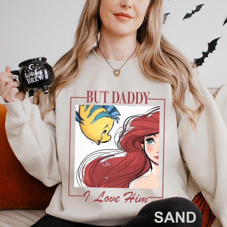 May include: Sand-coloured sweatshirt with a graphic of Ariel and Flounder from The Little Mermaid, and the text "BUT DADDY - I Love Him." The sweatshirt has a crew neck and long sleeves. The word "SAND" is printed at the bottom.