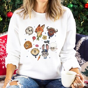 K&ouml;nnte beinhalten: Wei&szlig;es Sweatshirt mit farbenfrohen Cartoon-Illustrationen von Charakteren aus dem Film Zootopia. Das Design zeigt ein Faultier, einen Fuchs, einen L&ouml;wen, einen Geparden, ein Kaninchen, ein Schaf und einen B&uuml;ffel. Das Wort "WHITE" ist auf der Tasse gedruckt.