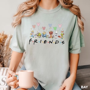 May include: A light green t-shirt featuring the word "FRIENDS" in a colorful font. Above the word are cartoon characters holding Mickey Mouse-shaped balloons. The shirt is a soft, comfortable material.